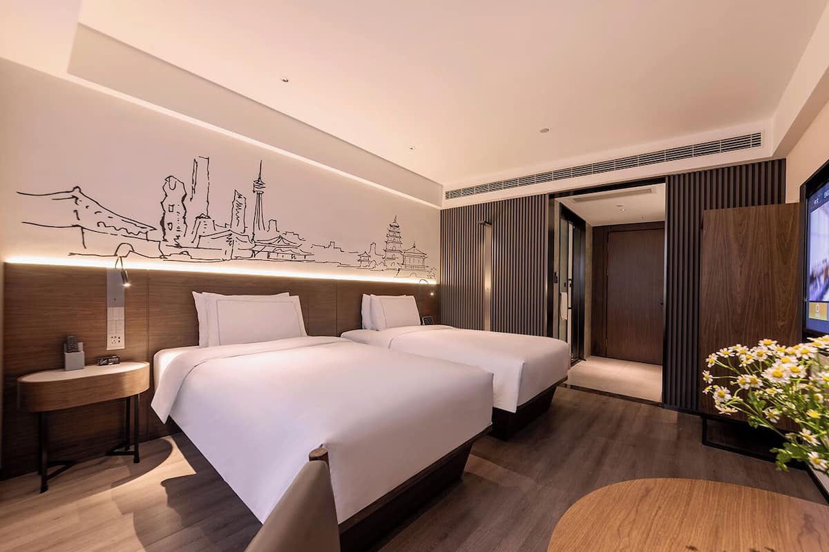Una habitación de hotel moderna con dos camas, un mural de paisaje urbano en la pared, una mesa redonda de madera y un televisor a la derecha.