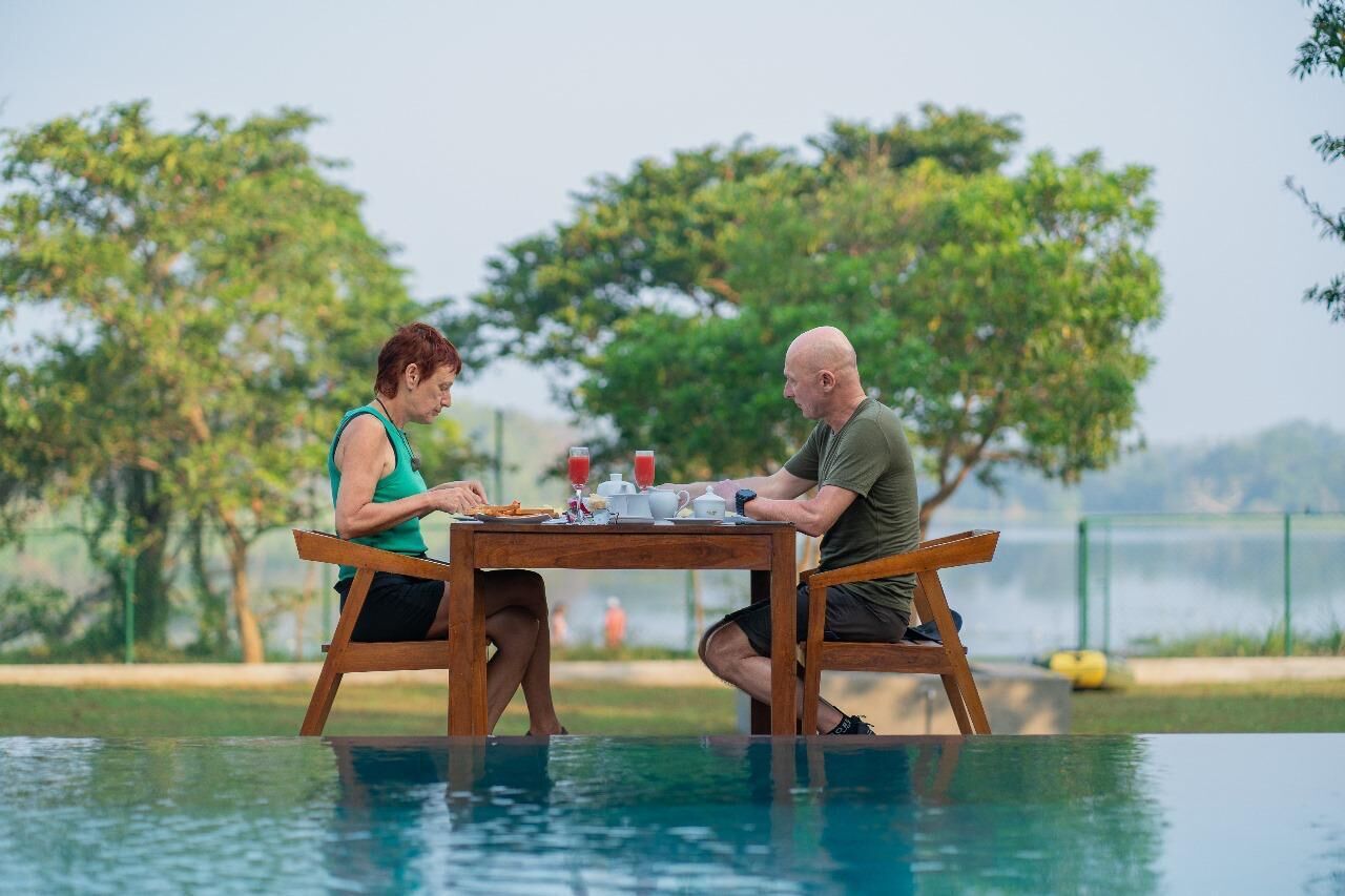 Two people dining at a wooden table by a poolside with a view of trees and a body of water in the background.