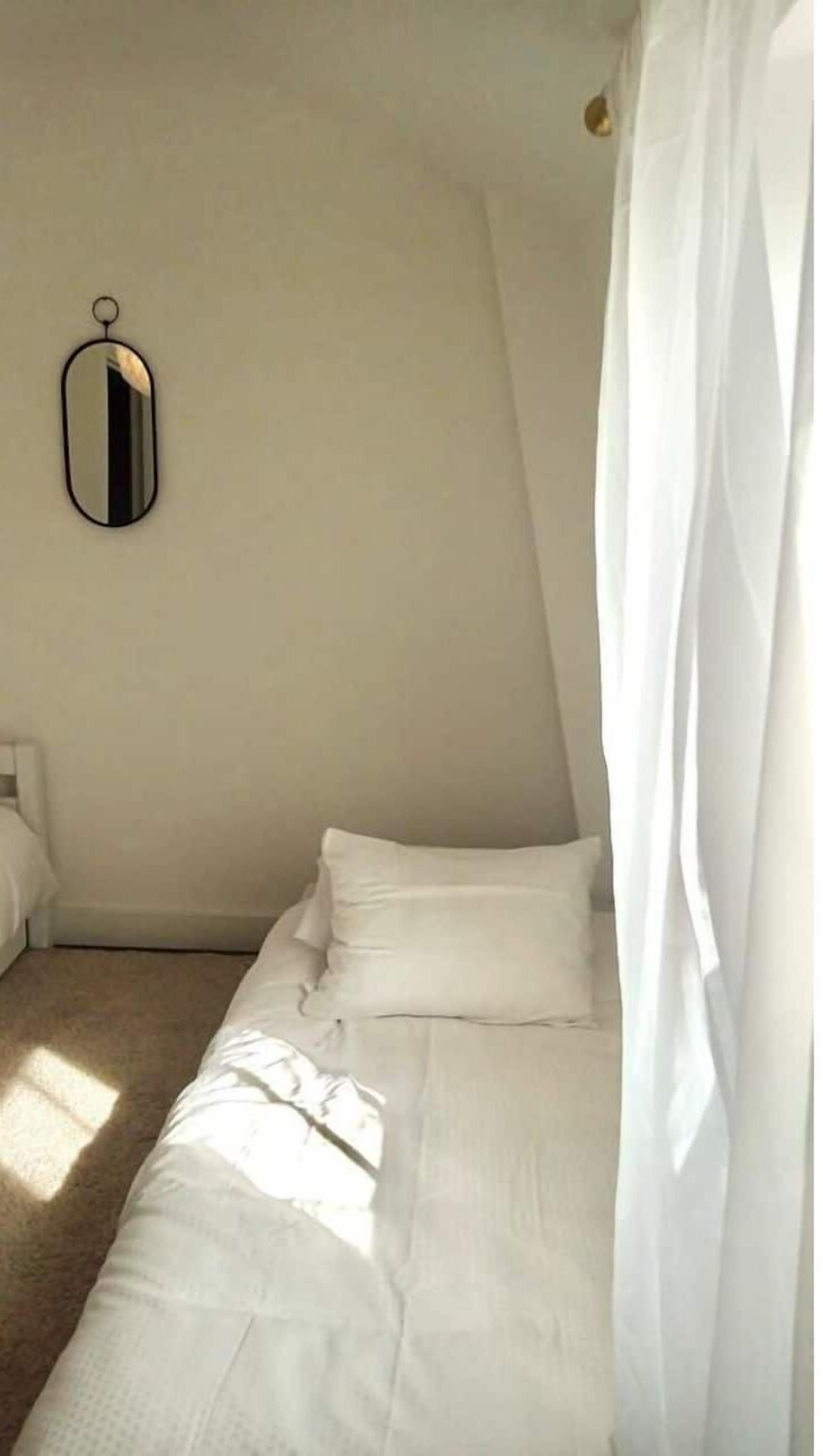 A bedroom with a single bed, white bedding, a hanging mirror, and a sheer curtain.