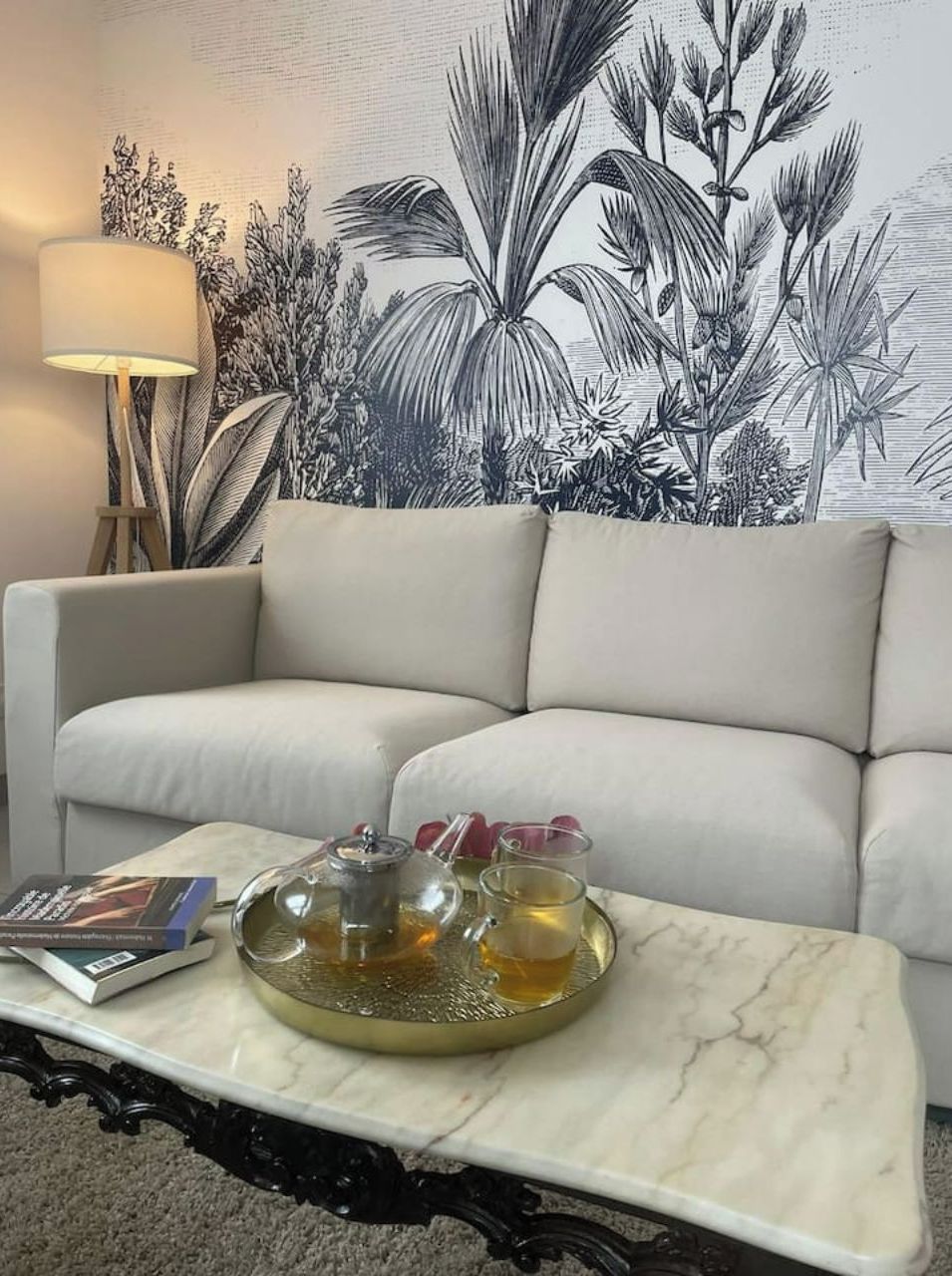 A living room with a white sofa, a marble coffee table with a teapot and cups, a lamp, and a wall with a tropical plant mural.