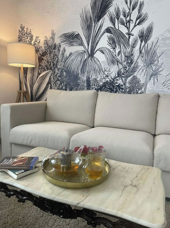 Un soggiorno con un divano bianco, un tavolino in marmo con teiera e tazze, una lampada e una parete decorata con un murale di piante tropicali.