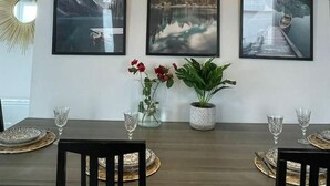 Un comedor con una mesa de madera dispuesta para dos comensales, tres cuadros de paisajes en la pared, una lámpara colgante de fibras tejidas y una planta en maceta.