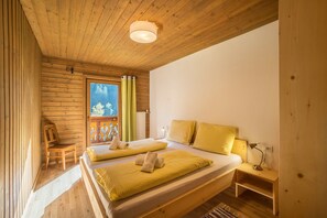 A wooden cabin bedroom with two beds, a chair, a nightstand, and a window with curtains.