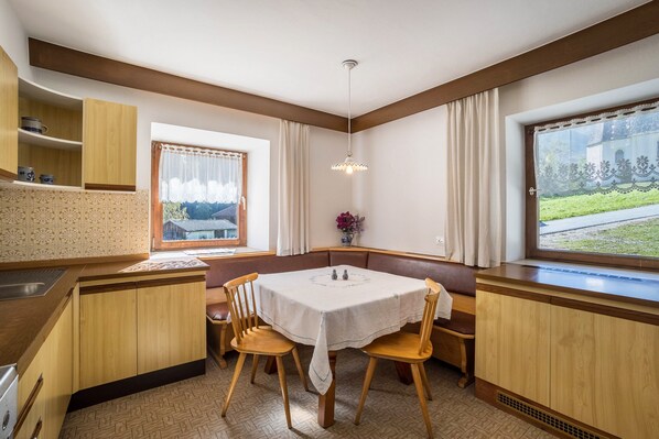 A kitchen with wooden cabinets, a dining table with chairs, a window with curtains, and a hanging light fixture.