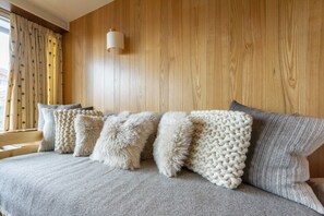 Uma cama com cobertor cinzento e várias almofadas, parede de madeira e janela com cortinas.