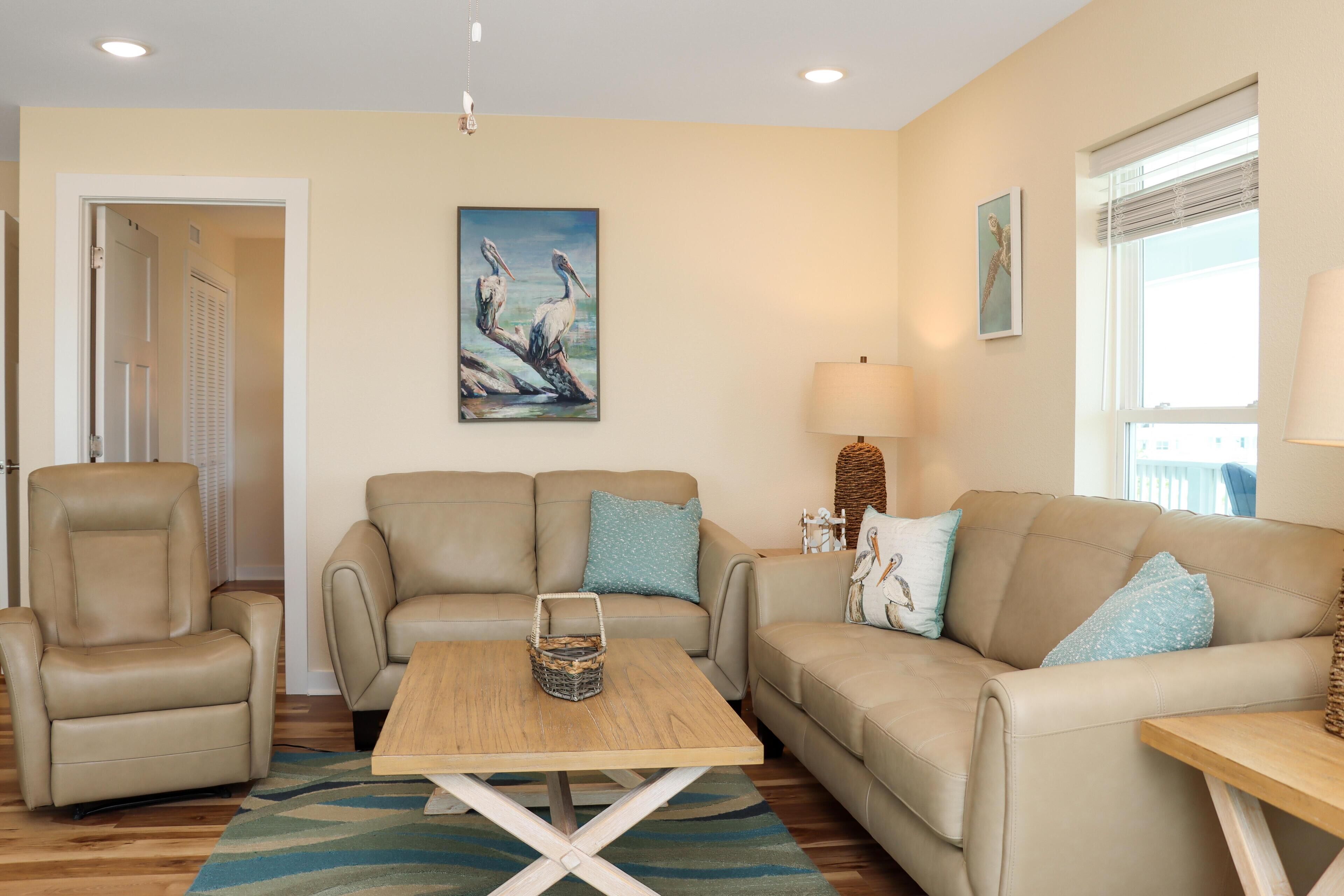 A living room with a beige sofa, a recliner, a wooden coffee table, and a painting of pelicans.