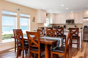 A dining area with wooden chairs and a table, a kitchen with white cabinets and stainless steel appliances, and a view of the ocean through large windows.