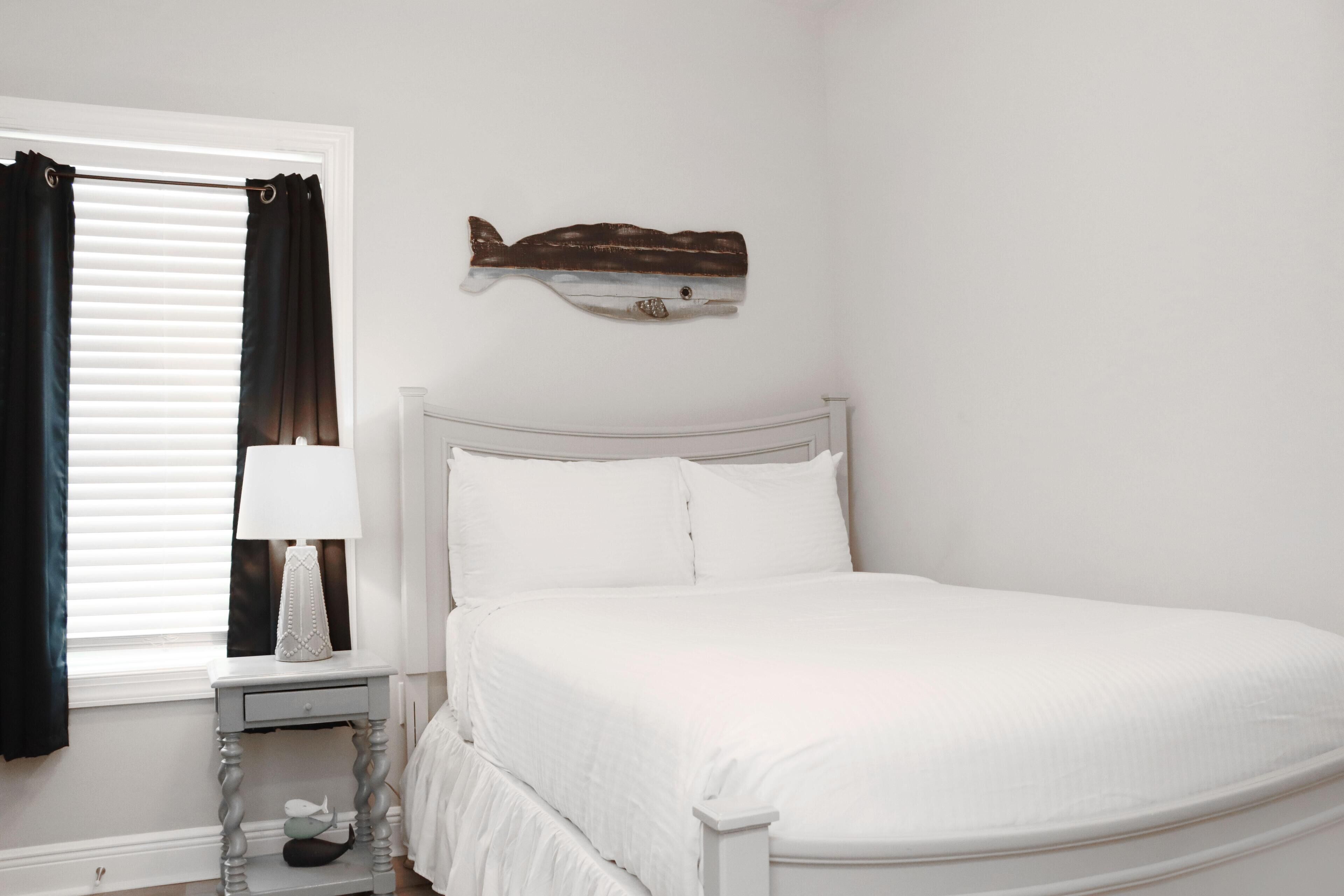 A bedroom with a white bed, a bedside table with a lamp, a window with blinds, and a wall-mounted fish decoration.
