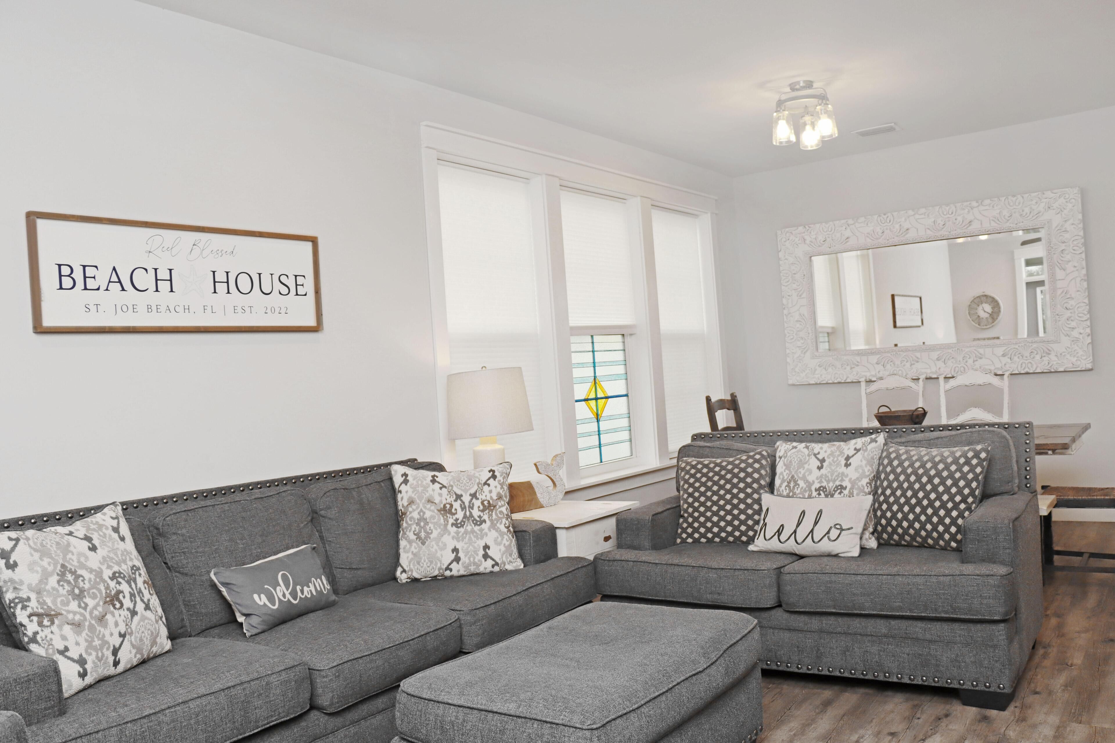 Ein Wohnzimmer mit einem grauen Ecksofa, einem Couchtisch, einem Beistelltisch, einer Lampe und einem gerahmten Schild mit der Aufschrift 'Beach House St. Joe Beach, FL | Gegründet 2022'.