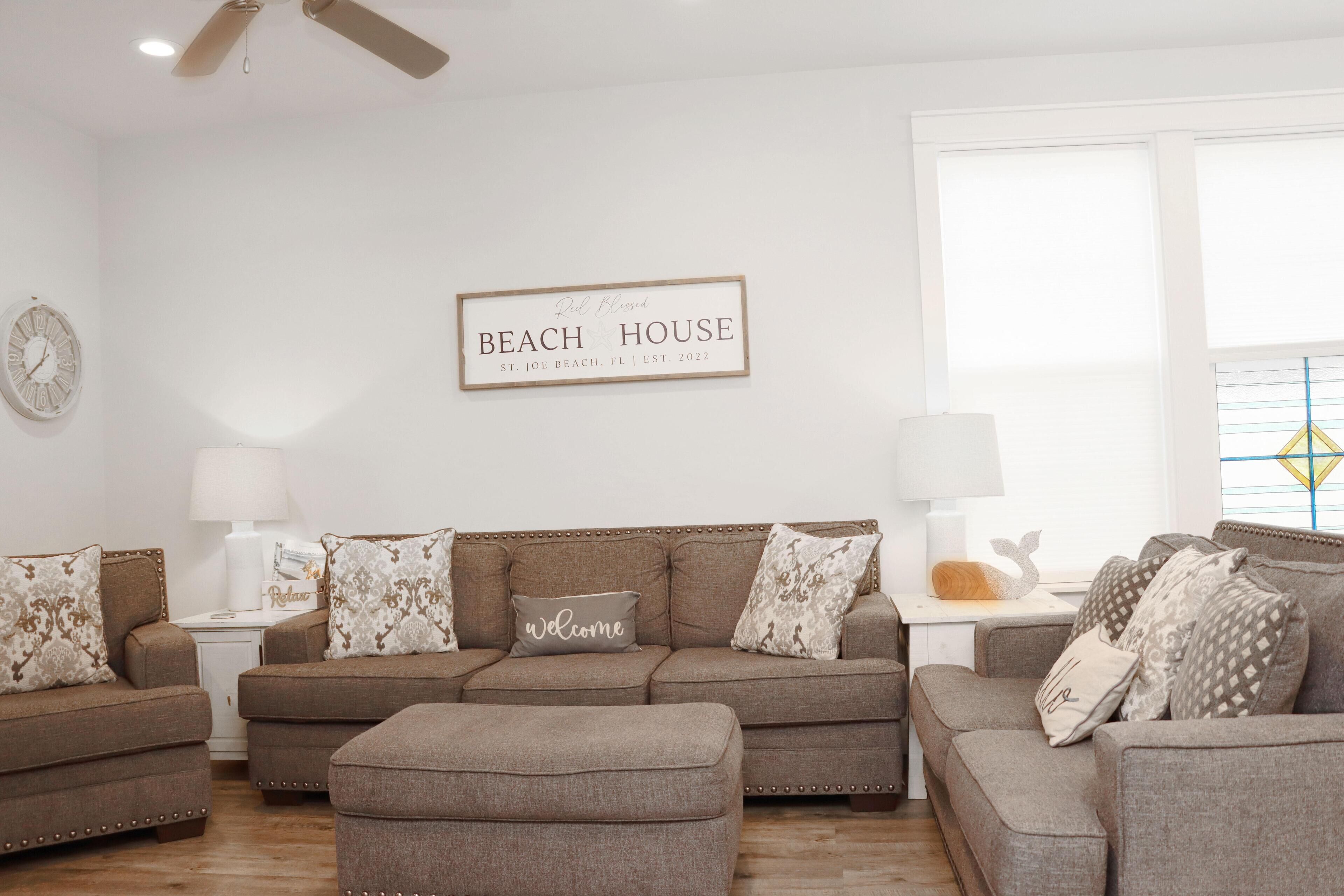 Ein Wohnzimmer mit einem beigefarbenen Sofagarnitur, einem Couchtisch und einem an der Wand befestigten Schild mit der Aufschrift 'Beach House St. Joe Beach, FL | Gegr. 2022'.