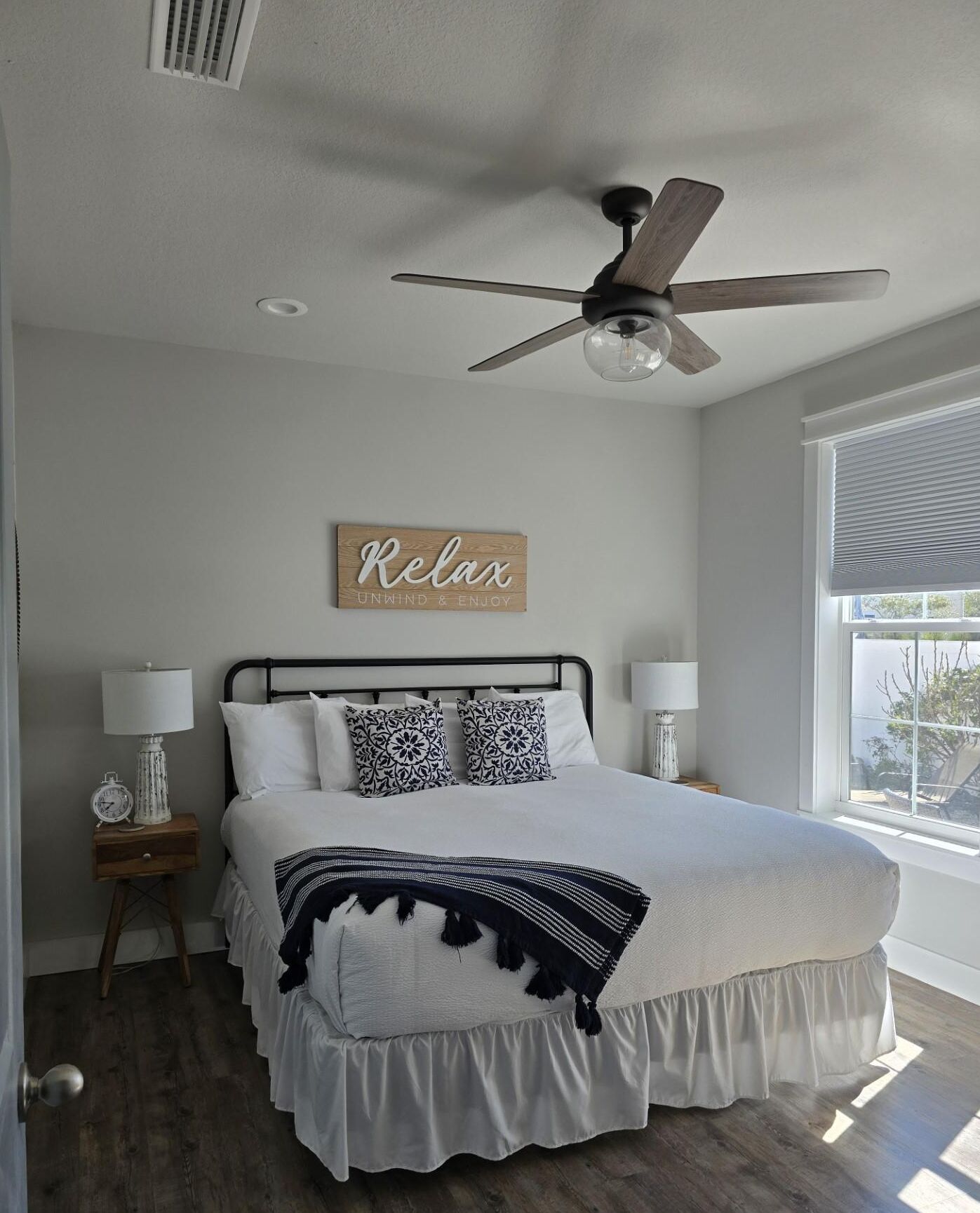 Une chambre à coucher avec un lit, un ventilateur de plafond, une fenêtre avec des stores et un panneau fixé au mur.