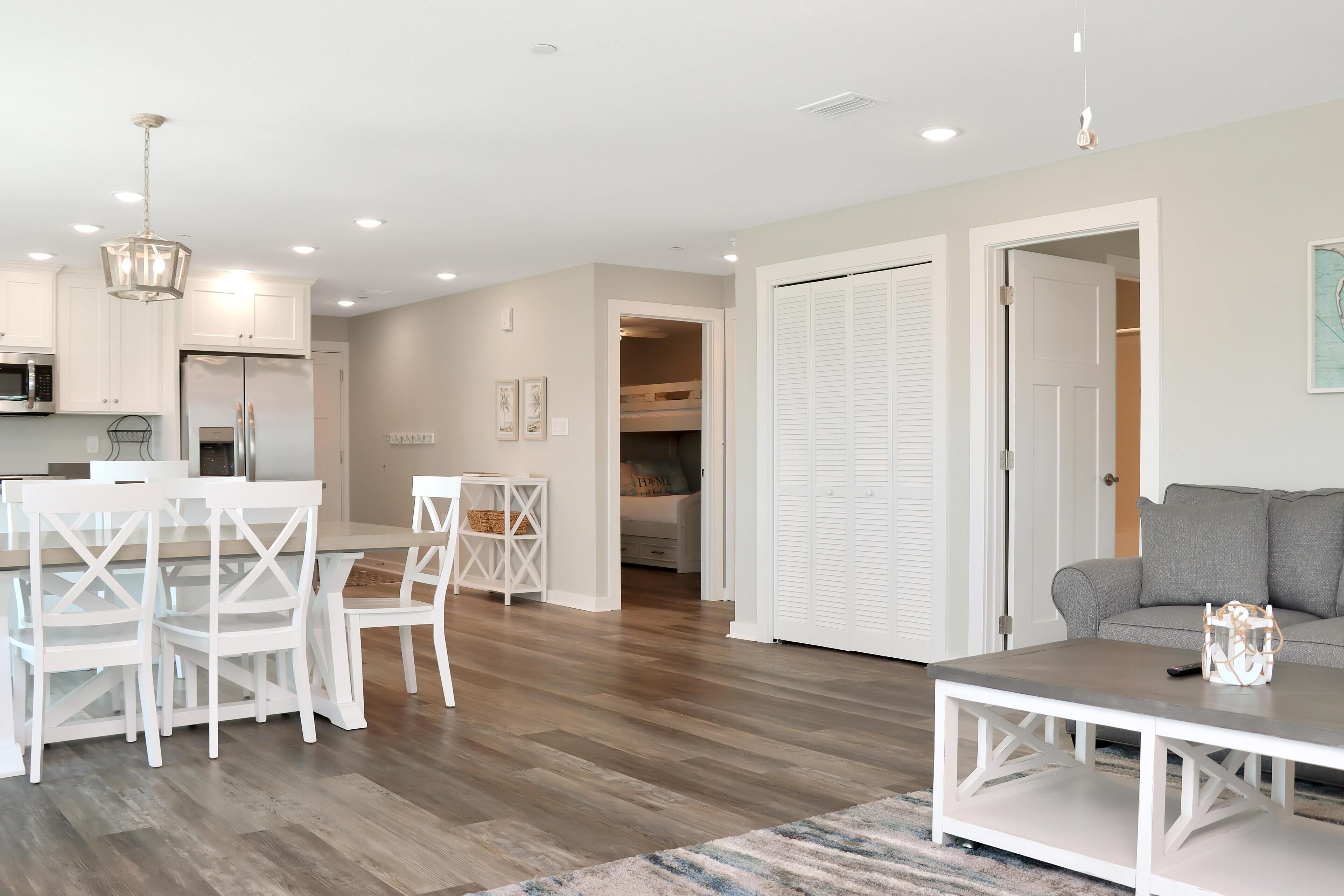 A modern kitchen with white cabinetry, a dining area with white chairs, and a living room with a grey sofa.