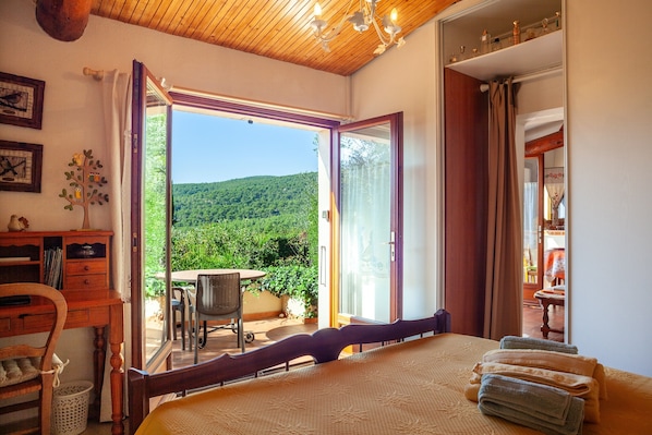 A bedroom with a bed, a desk, a chair, and a view of a balcony with a table and chairs.