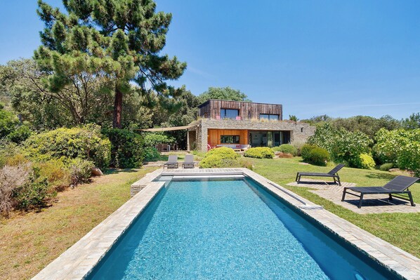 Une maison moderne dotée d’une piscine, de deux chaises longues et d’un jardin bien entretenu.