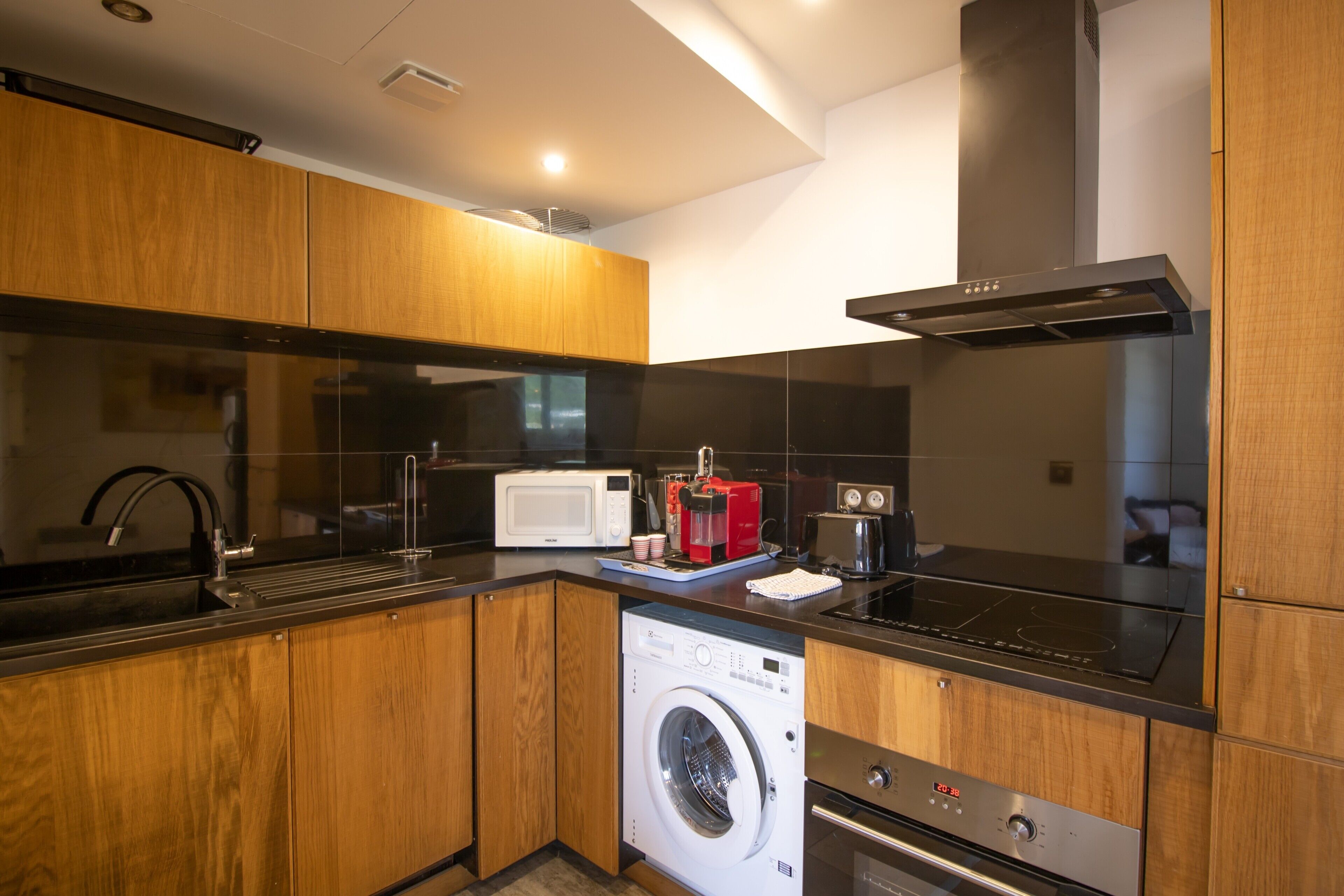 Una cocina moderna con armarios de madera, un lavamanos de acero inoxidable, un microondas, una cafetera, una lavadora y una estufa.