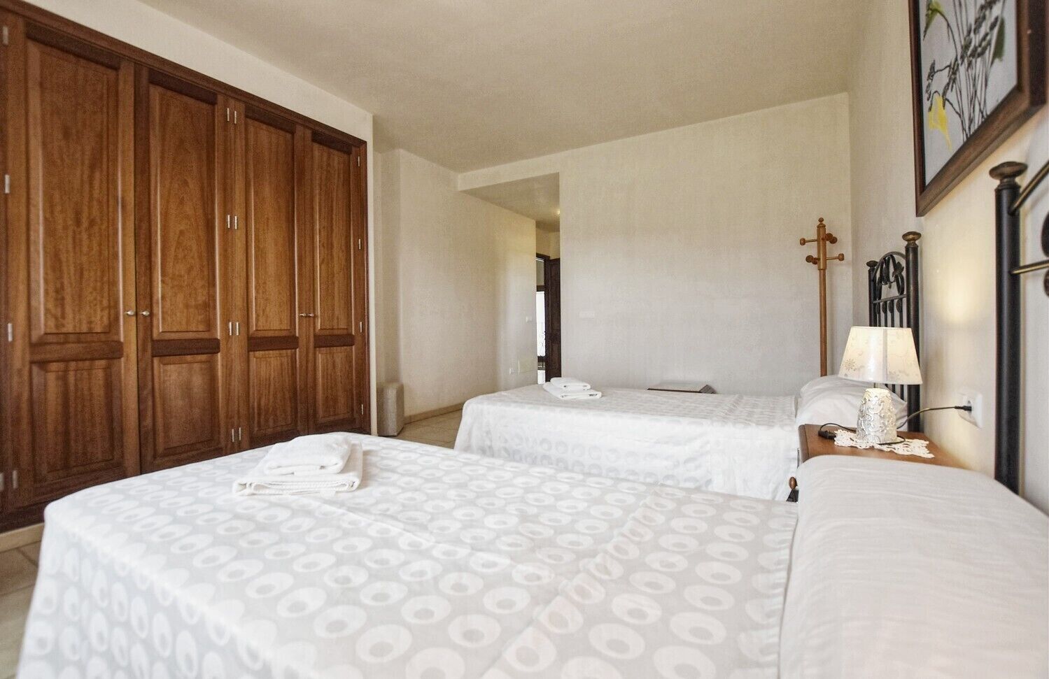 A hotel room with two beds, a wardrobe, a nightstand with a lamp, and a framed picture on the wall.