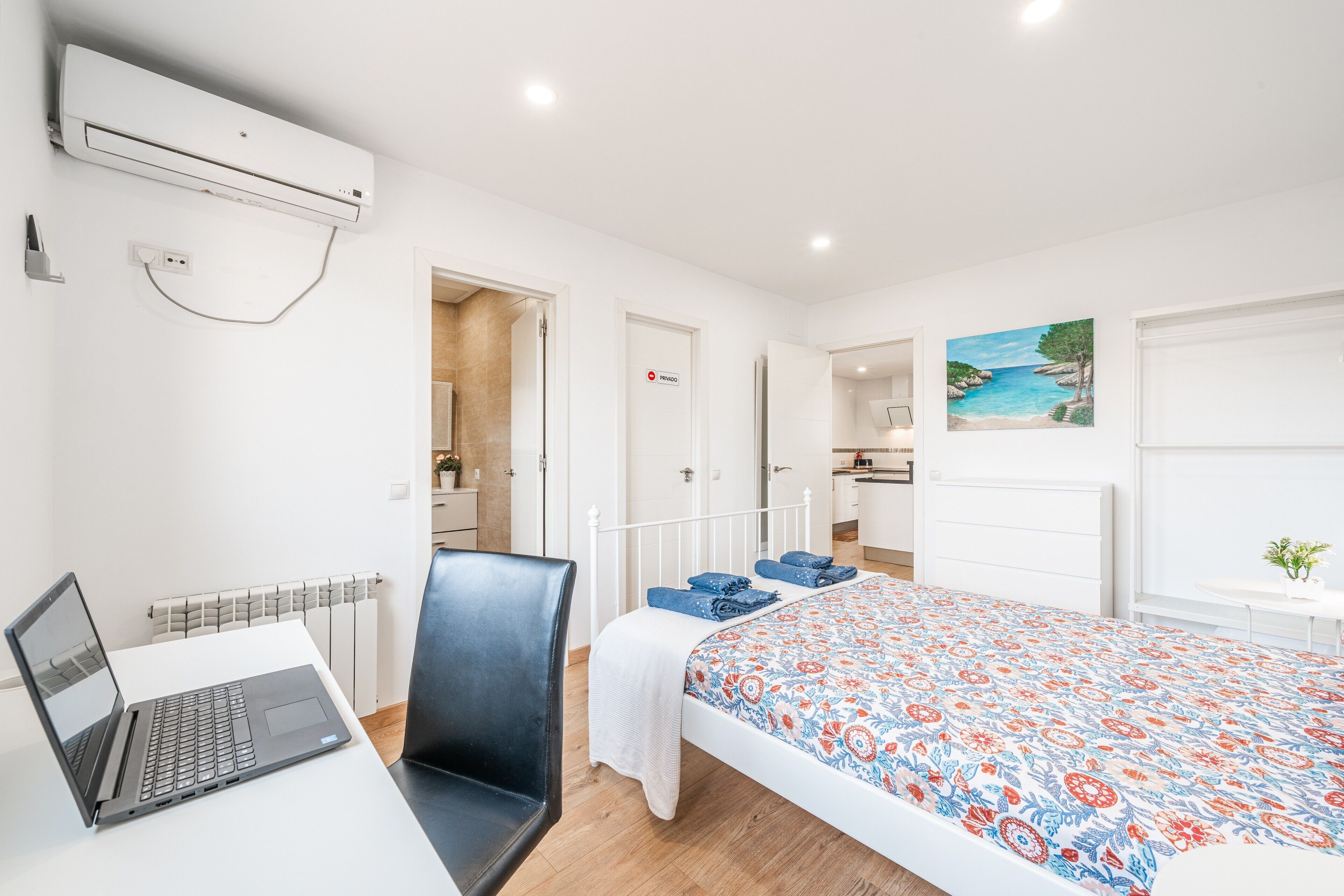 A modern hotel room with a bed, a desk with a laptop, a chair, a radiator, and a painting on the wall.