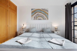 A bedroom with a bed, two bedside tables, a nightstand, a wooden wardrobe, a window with curtains, and a wall-mounted abstract painting.