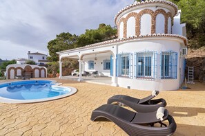 Ein Poolbereich mit Liegestühlen, einer überdachten Terrasse und einem weißen Gebäude mit blauen Toren.