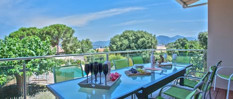 A glass-top table with green chairs on a balcony overlooking a scenic landscape with trees and mountains.