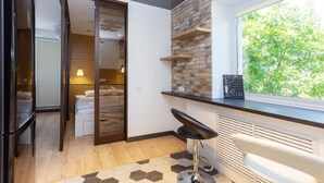Moderni hotellihuone, jossa on puulattia, suuri ikkuna, josta avautuu näkymä puita kohti, musta baarituoli ja liukuovi, joka johtaa kylpyhuoneeseen.