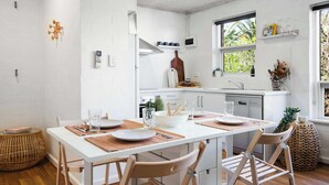 Uma cozinha moderna com armários brancos, uma mesa de jantar posta com pratos e talheres, e uma janela que permite entrada de luz natural.