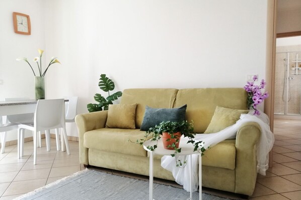 Una sala de estar con un sofá amarillo, una pequeña mesa blanca con una planta, y un comedor con sillas blancas.
