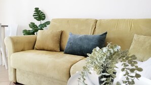 A beige sofa with three cushions, a potted plant, and a white side table in a room with white walls and a tiled floor.