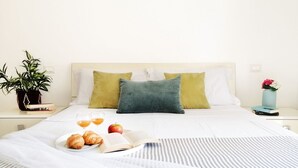 A neatly made bed with pillows, a bedside table with a plant, a book, and a plate with croissants and fruit.