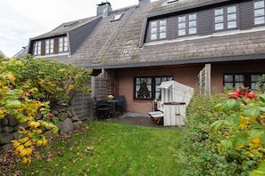 A two-story house with a sloped roof, a stone wall, and a small patio area with outdoor furniture.