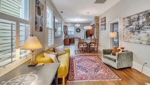 A spacious living area with a yellow sofa, a grey armchair, and a dining table. There is a large rug, a ceiling fan, and artwork on the walls.