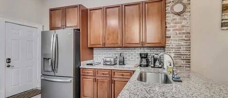 A modern kitchen with wooden cabinets, a stainless steel refrigerator, a granite countertop, and a brick accent wall.