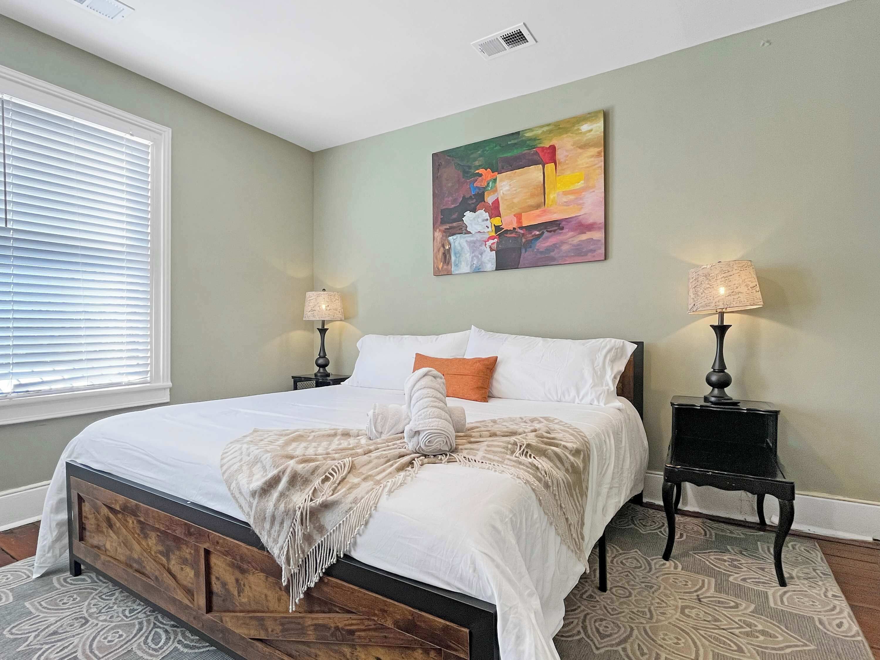 A bedroom with a large bed, two bedside tables with lamps, a window with blinds, and a colorful abstract painting on the wall.