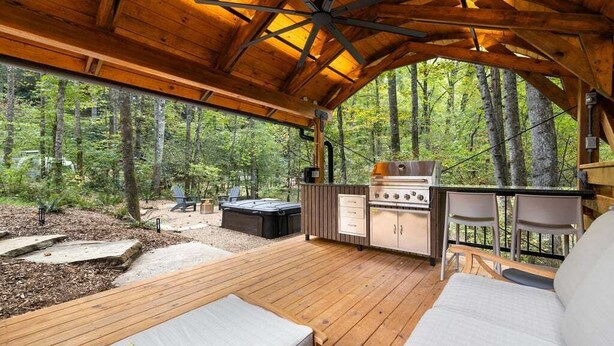 Een overdekt terras buiten met een barbecue, zitplaatsen en uitzicht op het bos.