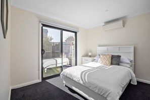 A bedroom with a bed, a nightstand, a lamp, a window with blinds, and an air conditioning unit.