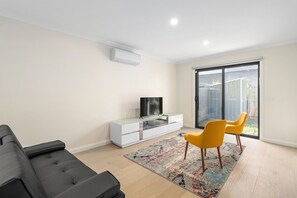 A modern living room with a sofa, a TV on a white cabinet, and a large sliding glass door to the outside.
