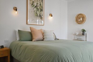 Una cama bien hecha con almohadas, una mesita de noche con una lámpara y un estante en la pared con una planta.