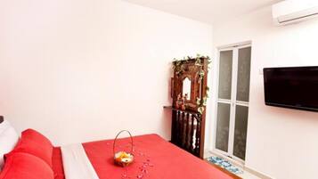 A bedroom with a red bedspread, a wooden cabinet with greenery, a television on the wall, and a window with a view.