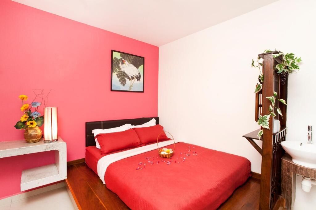 Ein Schlafzimmer mit roter Bettdecke, einem gerahmten Bild an der Wand, einem Nachttisch mit Blumenvase und einem Holzpflanzentisch mit Grünpflanzen.