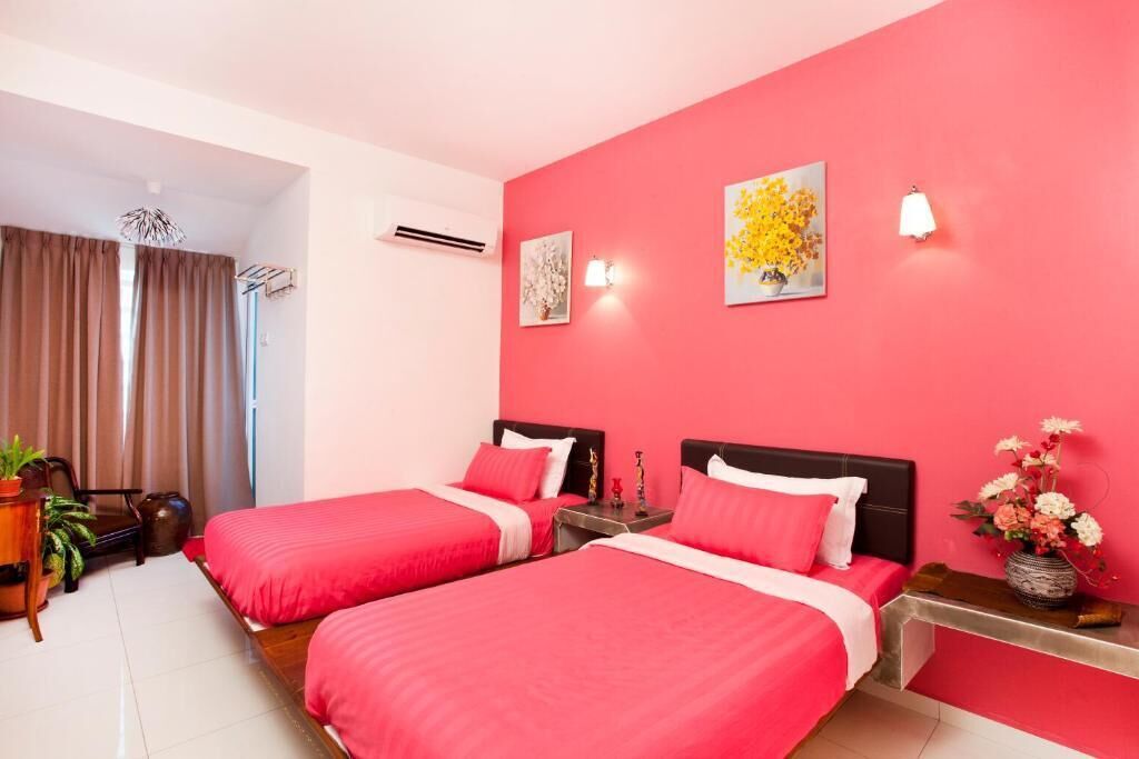 A hotel room with two beds, a red accent wall, a window with curtains, and a small table with a vase of flowers.