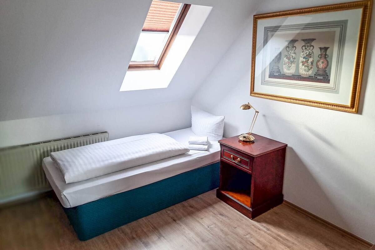 A small, neatly arranged bedroom with a single bed, a radiator, a small table with a lamp, and a framed picture on the wall.