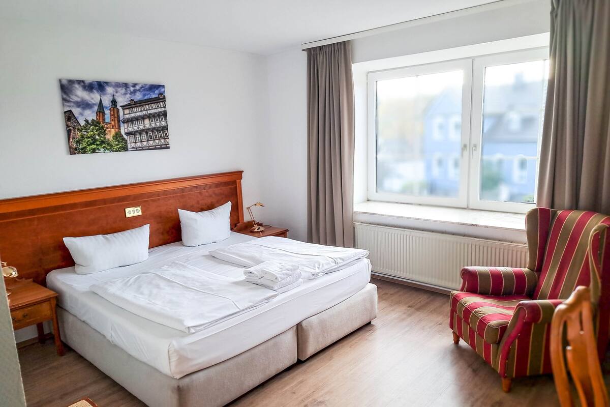 A hotel room with a large bed, a wooden headboard, a window with curtains, a striped armchair, and a painting on the wall.