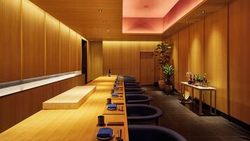 Una larga mesa de madera con servilletas azules y cubiertos, rodeada de sillas oscuras en una habitación moderna con paredes de madera y una lámpara en el techo.
