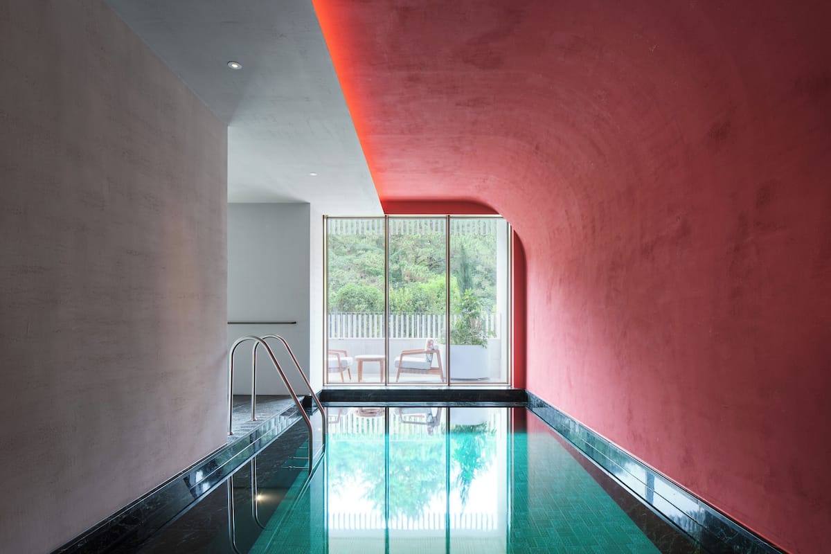 A modern indoor swimming pool with a glass wall, a red accent wall, and a view of greenery outside.