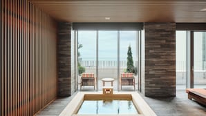 A modern spa area with a wooden floor, a rectangular hot tub, two chairs, and a large glass window offering a view of the sea. - Ananti At Busan Village (Busan)
