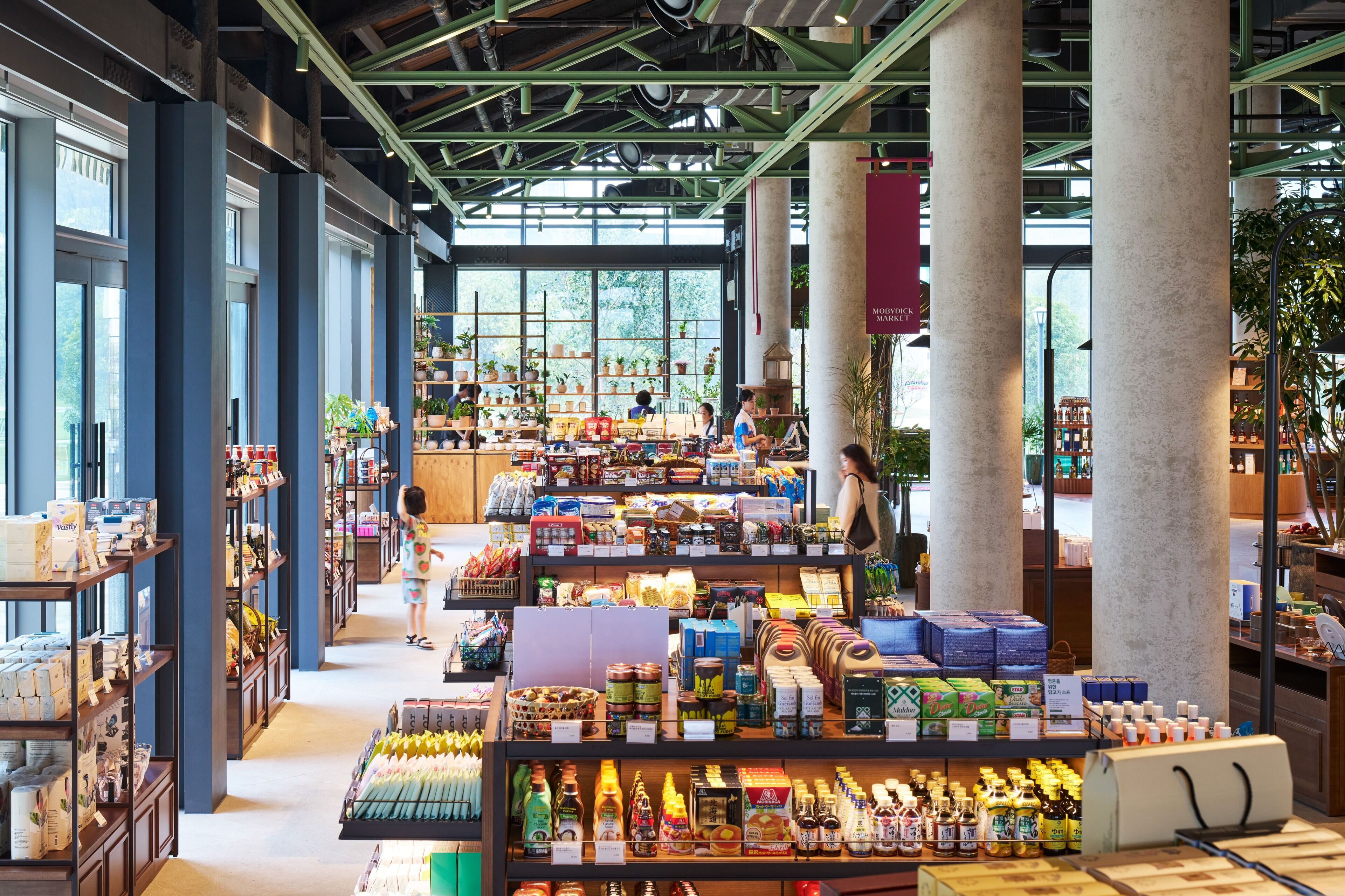 A spacious indoor market with wide aisles, wooden shelves stocked with various goods, and large windows allowing natural light.