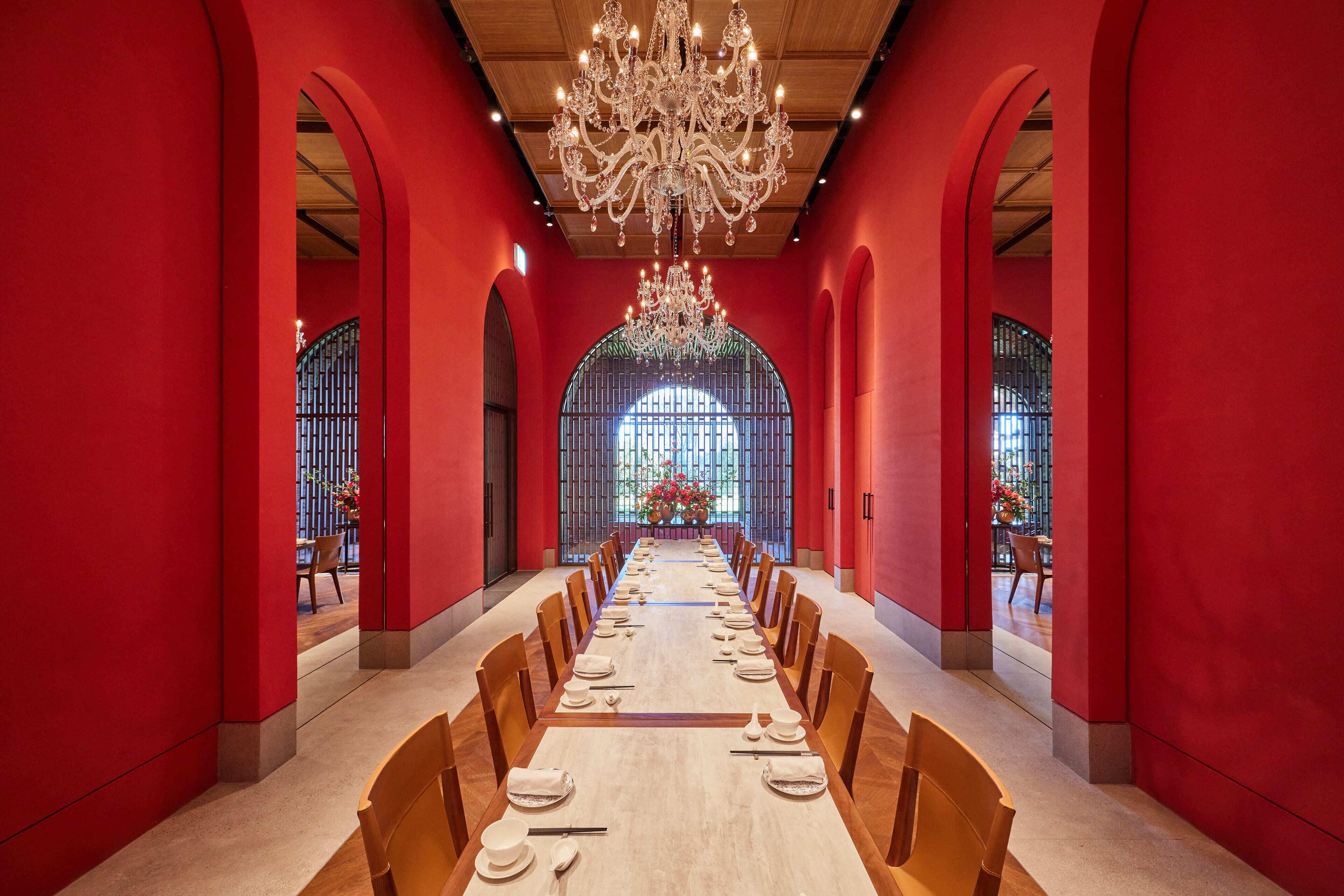 Une longue table à manger, dressée pour un repas, se trouve dans une pièce aux murs rouges et ornée d’un grand lustre.