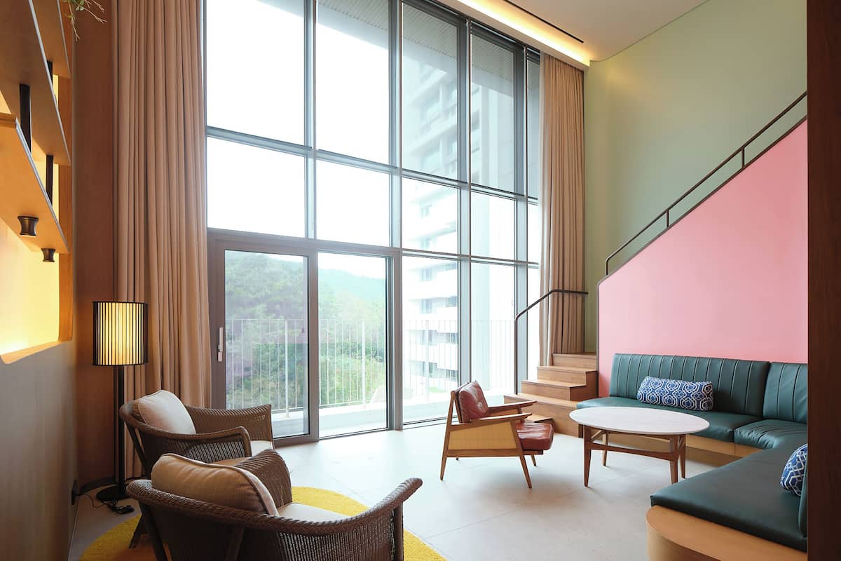 A modern hotel room with large windows, a green sofa, wicker chairs, a small table, and a staircase with a pink wall.