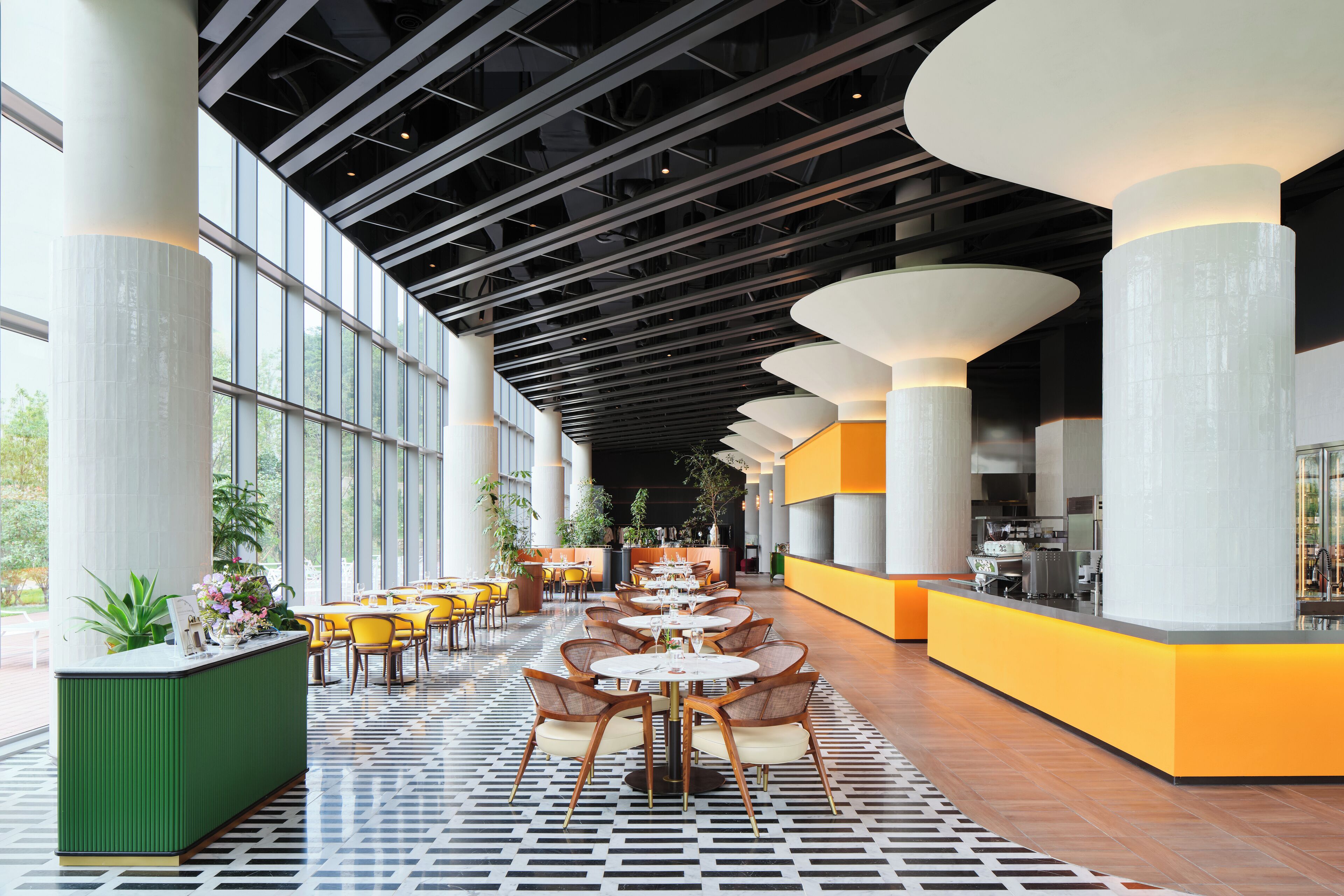 A spacious indoor area with a long row of tables and chairs, large windows, and a modern ceiling design.