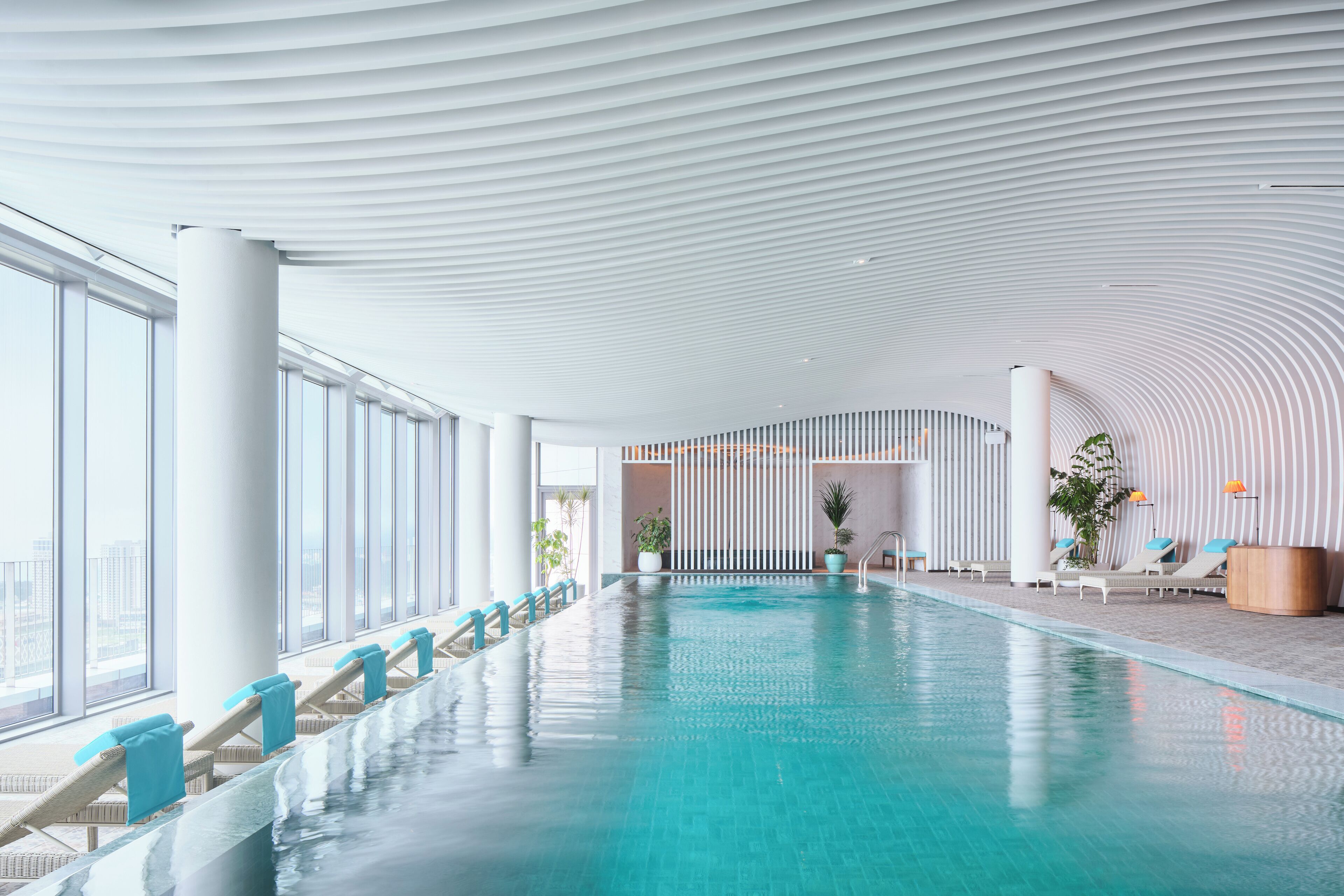 A modern indoor swimming pool with a curved ceiling, large windows, and lounge chairs along the poolside.